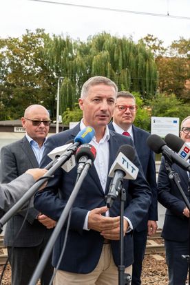 Zdjęcie przedstawia mężczyznę, który mówi do mikrofonów. W tle za nim stoi trójka mężczyzn.