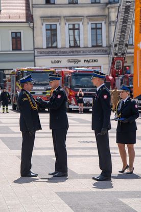 Zdjęcie przedstawia trzech mężczyzn i jedną kobietę w mundurach strażackich. W tle ludzie i wozy strażackie.