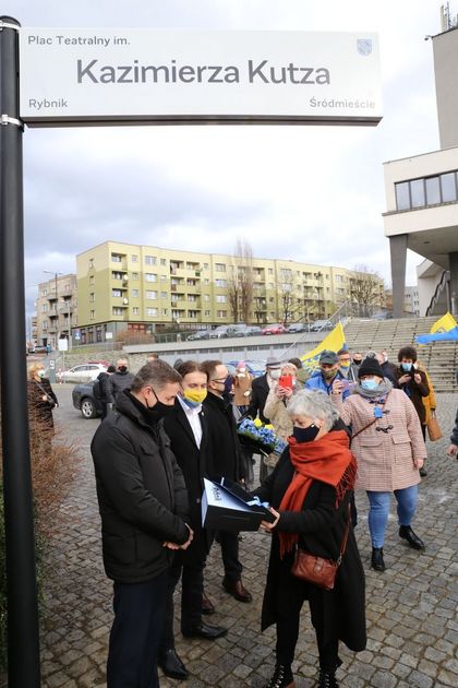 Kilkanaście ubranych zimowo osób na otwartej przestrzeni. Kilka osób stoi pod znakiem informującym, o nazwie placu, o którym mowa w tekście.