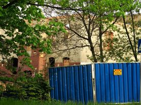 Swierklanska fot.LucynaTyl 12 Na pierwszym planie zielone drzewa i niebieski płot blaszany. W tle zrujnowany, opuszczony budynek w czasie gruntownego remontu.