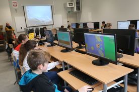 Klasa szkolna, w ławkach siedzą dzieci przed monitorami komputerów