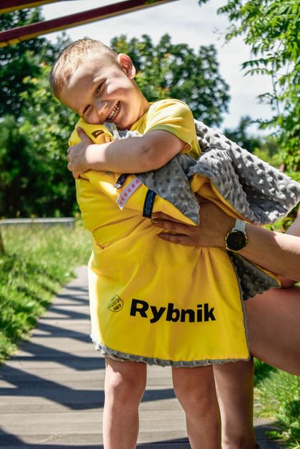 Kilkuletni chłopczyk otulony żółto-szarym kocykiem z napisem Rybnik i herbem miasta. 