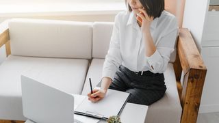 Kobieta siedząca na kanapie, trzymająca w jednej ręce telefon, drugą robi notatki na kartce. Przed nią stoi laptop 