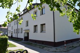 Jasny kilkupiętrowy budynek mieszkalny, na pierwszym planie zieleń i osiedlowy chodnik, w tle niebo. 