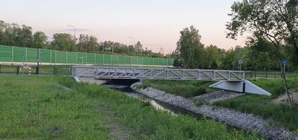 Wijąca się wśród zieleni rzeka z mostkiem dla rowerzystów i pieszych. W tle zielone ekrany akustyczne przy drodze szybkiego ruchu.