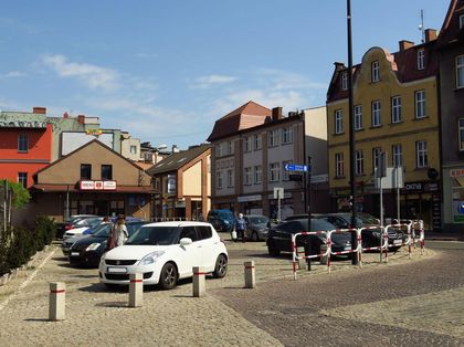 Wybrukowany placyk w centrum miasta. Na nim zaparkowane samochody. Wokół kamienice. 