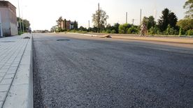 Droga w trakcie remontu, świeżo położony asfalt. Wzdłuż drogi domy, zieleń.