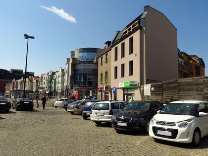 Wybrukowany placyk w centrum miasta. Na nim zaparkowane samochody. Wokół kamienice. 