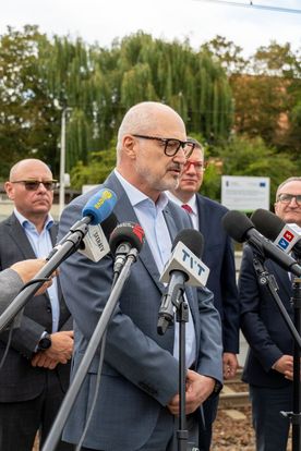 Zdjęcie przedstawia mężczyznę mówiącego do mikrofonu. W tle widać trzech mężczyzn.