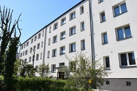 Jasny kilkupiętrowy budynek mieszkalny, na pierwszym planie zieleń, w tle błękitne niebo. 