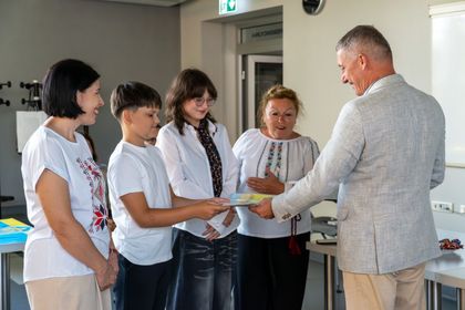 Zdjęcie przedstawia trójkę grupę osób, które rozmawiają. Chłopiec przekazuje mężczyźnie prezent. 