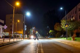 Widok ulicy w nocy. Na środku znajduje się osoba, która przechodzi przez przejście dla pieszych. W tle widać samochód w ruchu.