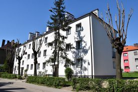 Jasny kilkupiętrowy budynek mieszkalny, na pierwszym planie zieleń, w tle błękitne niebo i inne zabudowwania. 