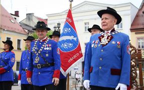 Grupa mężczyzna w strojach Bractwa Kurkowego. W tle sztandar
