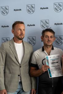 Zdjęcie przedstawia dwóch mężczyzn pozujących do zdjęcia. Jeden z nich trzyma dyplom.