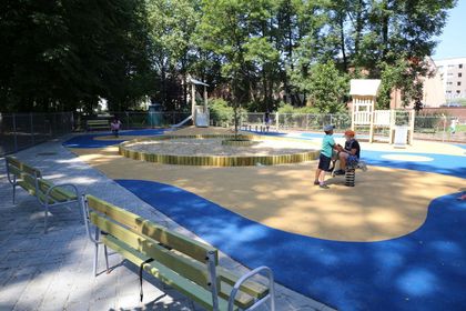 Plenerowy plac zabaw, w otoczeniu zieleni parkowej. Piaskownica, drwniana jeżdżalnia, huśtawki, bawiące się dzieci. 