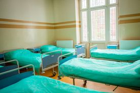 Jasna sala szpitalna z oknem, po obu stronach rząd zasłanych łóżek z zieloną pościelą 