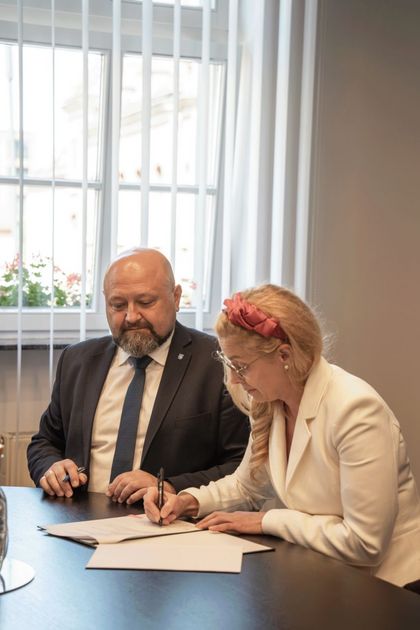 Zdjęcie przedstawia dwójkę osób siedzących przy stole na oficjalnym spotkaniu.