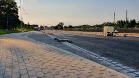 Droga w trakcie remontu, świeżo położony asfalt. Wzdłuż drogi domy, zieleń.