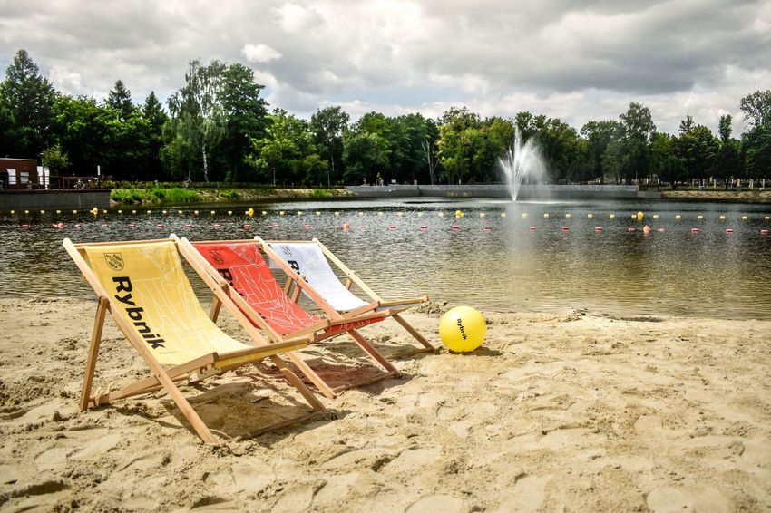 Kąpielisko z fontanną na środku. Na pierwszym planie plaża, a na niej trzy kolorowe leżaki z napisem Rybnik. 