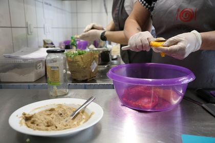 Dwie osoby pracują przy przygotowywaniu posiłku, naczynia z produktami spożywczymi na metalowym kuchennym blacie.