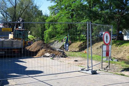 Roboty budowlane na drodze wzdłuż rzeki. W tle most, wokół drzewa i zabudowa miejska. 