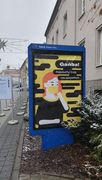 Zdjęcie stojącej przy śródmiejskiej ulicy gabloty informacyjnej. W gablocie plakat z napisem Gańba