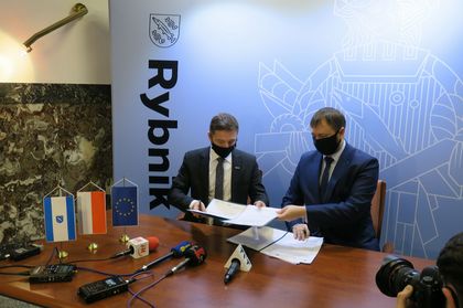 Dwóch mężczyzn podpisujących dokumenty, przy stole, na tle ścianki z napisem Rybnik. 