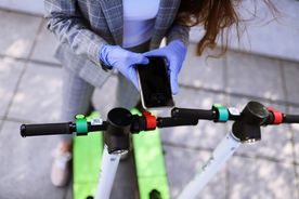 Fragment postaci elegancko ubranej kobiety w niebieskich rękawiczkach trzymającej w rękach smartfona. Kobieta podtrzymuje zielono-czarną hulajnogę. 