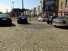 Wybrukowany placyk w centrum miasta. Na nim zaparkowane samochody. Wokół kamienice. 