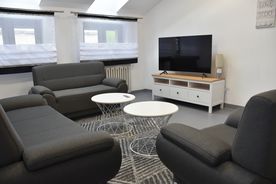 Jasne wnętrze, szara sofa i fotele, dwa małe białe stoliki, telewizor na białej szafce.