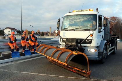 Ulice miasta, na nich pługopiaskarka i pracownicy służb komunalnych z szuflami do odśnieżania.