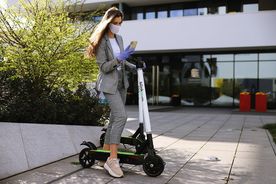 Elegancko ubrana kobieta w niebieskich rękawiczkach i maseczce ochronnej trzyma w jednej ręce smartfona, a drugą podtrzymuje hulajnogę. W tle zieleń i biurowiec. 