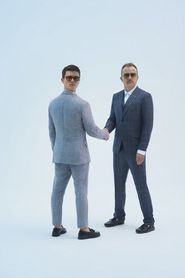 Karaś i Rogucki. Dwóch mężczyzn w okularach i garniturach w niebieskiej tonacji na jasnym tle. Mężczyźni trzymają się za ręce, głowa jednego z nich jest przekręcona o 180 stopni. 