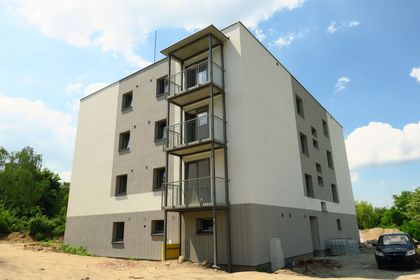 Jasny, kilkupiętrowy budynek mieszkalny z balkonami na tle błękitnego nieba z chmurami. Obok zieleń.