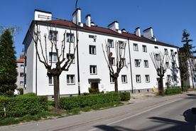 Jasny kilkupiętrowy budynek mieszkalny, na pierwszym planie zieleń oraz chodni i ulica, w tle błękitne niebo. 