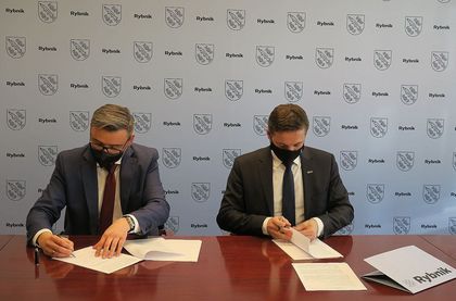 Prezydent Rybnika Piotr Kuczera i wójt Pilchowic Maciej Gogulla podpisują umowę przy stole konferenycyjnym. W tle ścianka reklamowa z herbem Rybnika