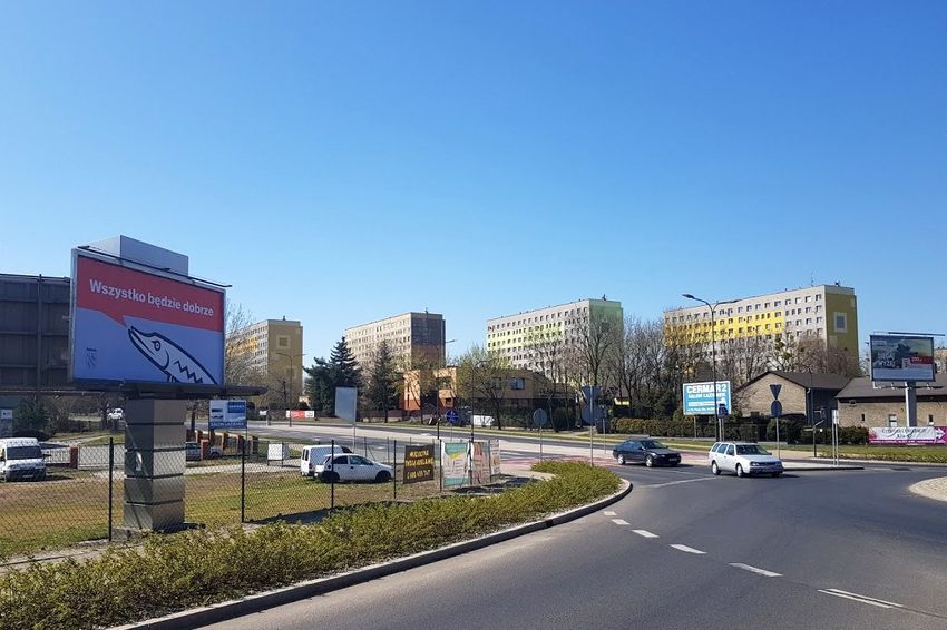 Zebrzydowicka 2 Billboard na tle błękitnego nieba, bloków. Z przodu ulica, w tle jadące samochody.