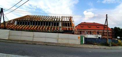 Droga, po drugiej stronie za płotem plac budowy, po prawej piętrowy, ceglany budynek pocysterskiego dworu w trakcie remontu. Po lewej budynek w trakcie remontu dachu - widoczna konstrukcja dachowa. W tle błękitne niebo z białymi chmurami. 