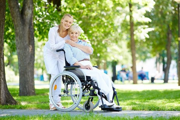Wiosenny park, dwie kobiety - starsza siedzi w wózku inwalidzkim, młodsza stoi i ją obejmuje. 