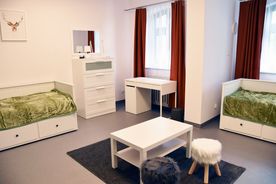 Pokój sypialny, jasne wnętrza, dwa łóżka, duże okna z zasłonami, na środku pokoju biały stolik.