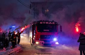 Wóz strażacki przejeżdżający zaśnieżoną drogą. Obok strażacy z pochodniami. 