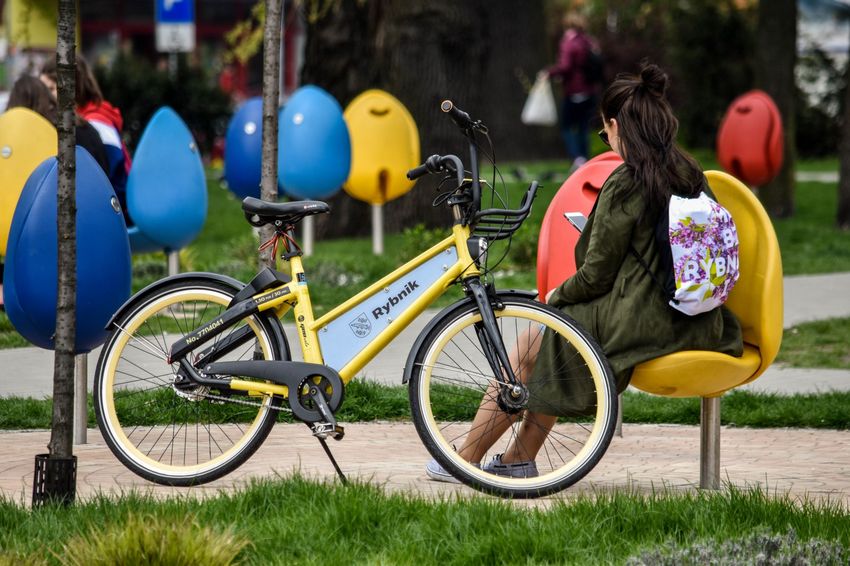 Rower miejski fotPR2 Żółty rower miejski stojący na skwerze. Na ławce kobieta siedząca tyłem