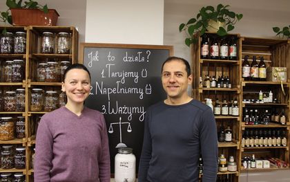 Zdjęcie przedstawia kobietę i mężczyznę, którzy pozują do zdjęcia. W tle sklepu, tablica i słoiki.