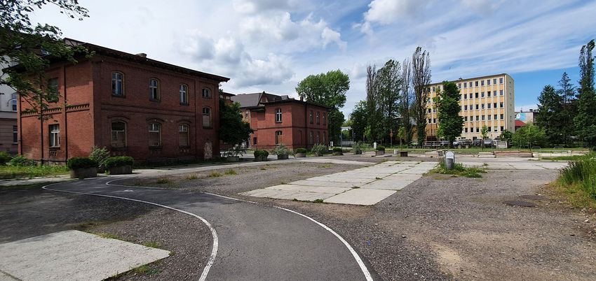 Ceglane budynki, na pierwszym planie plac, w tle inne zabudowania, zieleń i niebo.
