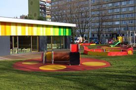 Parterowy budynek, pomalowany w kolorach tęczy. W tle wieżowiec mieszkalny, na pierwszym planie plac zabaw. 