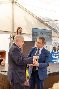 Na zdjęciu widać dwóch mężczyzn. W tle kobieta. Zdjęcie zrobione na oficjalnym wydarzeniu w pomieszczeniu.