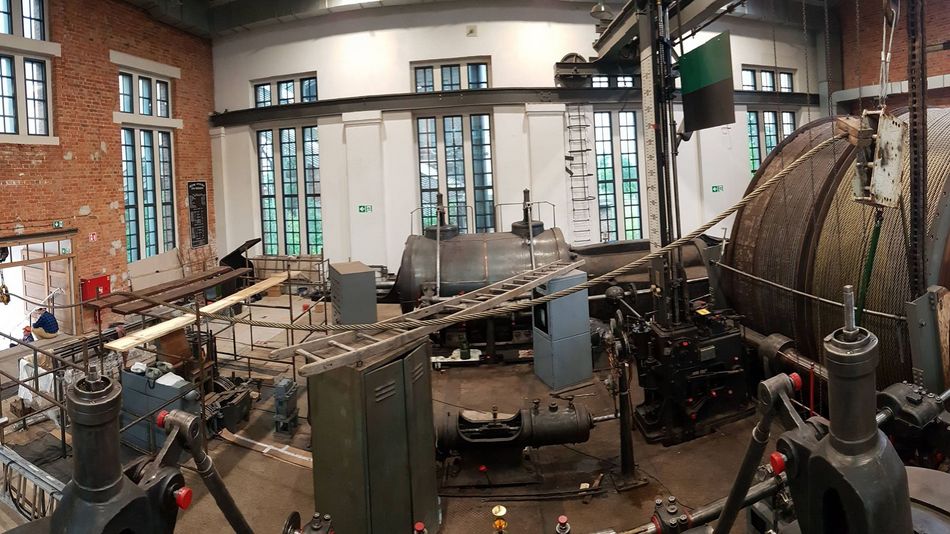 Wnętrze zabytkowej kopalni, przestronna hala z dużymi oknami, ceglanymi elementami na ścianach. Widoczne urządzenia maszyny parowej, w tym bęben z liną wyciągową.