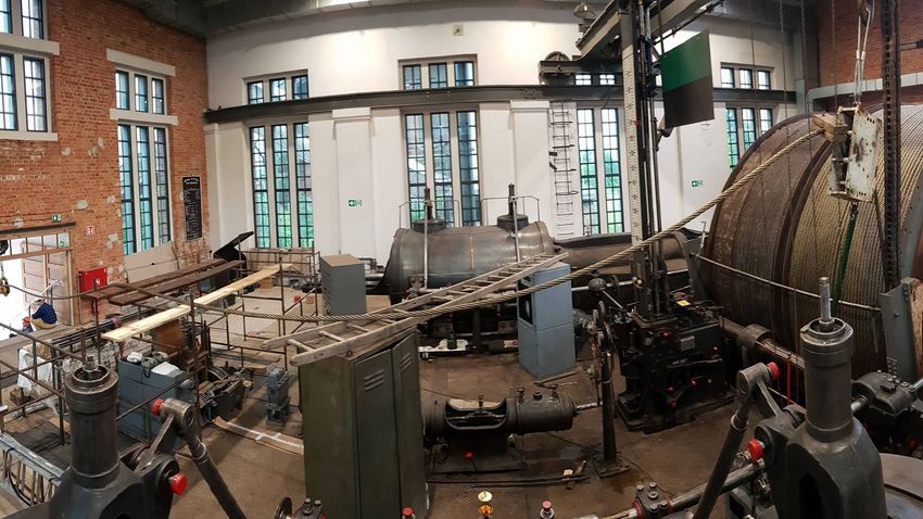 Wnętrze zabytkowej kopalni, przestronna hala z dużymi oknami, ceglanymi elementami na ścianach. Widoczne urządzenia maszyny parowej, w tym bęben z liną wyciągową.