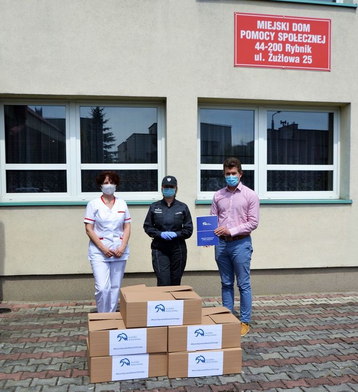 Maseczki SW Trzy osoby - pielęgniarka, funkcjonariuszka służby więziennej oraz mężczyzna stoją na tle budynku. Na pierwszym planie kartony z nadrukiem służba więzienna.